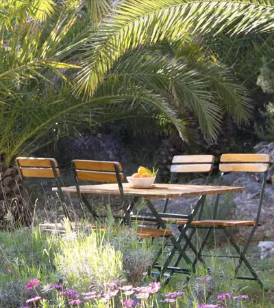 Wooden garden table and chairs under palm fronds with flowers in the garden of House Agave